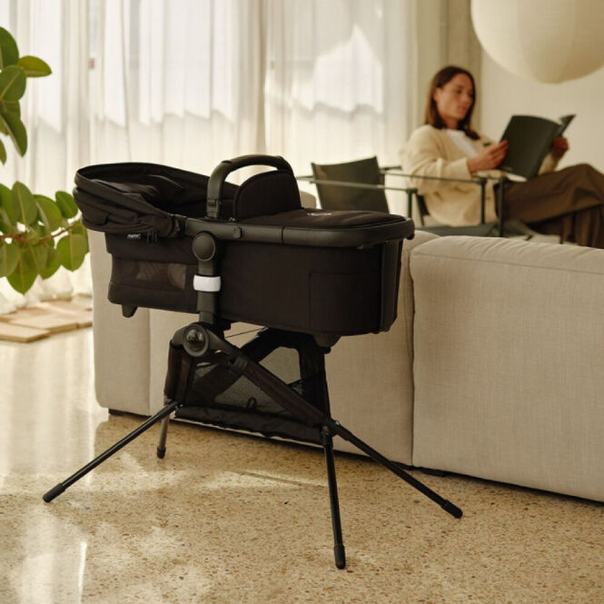 A Bugaboo bassinet placed on a bassinet stand, inside a cosy living room. In the background, a mom is reading and relaxing.