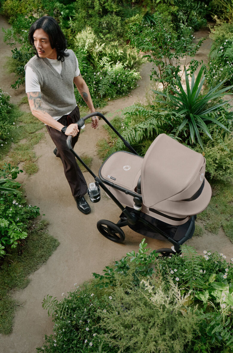 Ein Elternteil geht mit einem Bugaboo Fox 5 auf einem unbefestigten Weg in einem Park spazieren. Der Kinderwagen ist in Wüstentaupe gehalten.