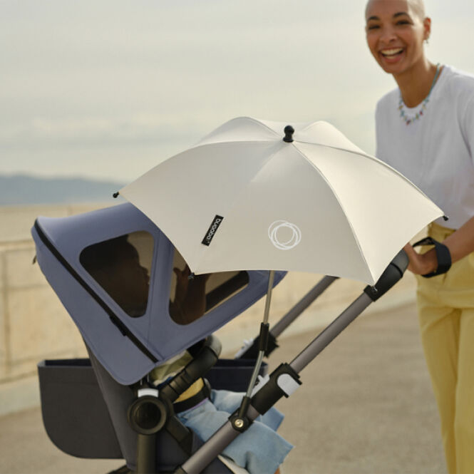 A mom takes her kid out for a walk in a sandy area. The pushchair is equipped with a blue breezy sun canopy and a white parasol.