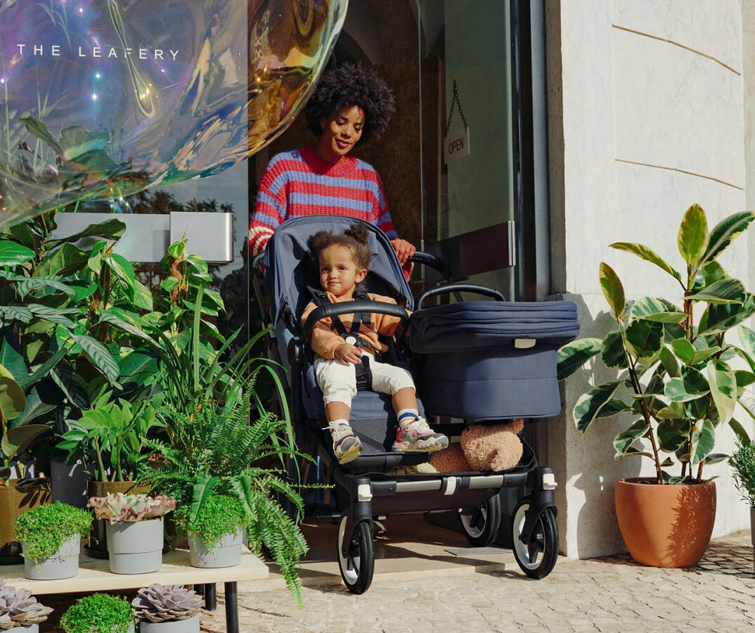 A mom exits a store, pushing a Bugaboo Donkey 5 Duo with a seat and a carrycot. A toddler sits in the seat. The store's entrance is surrounded with greenery.
