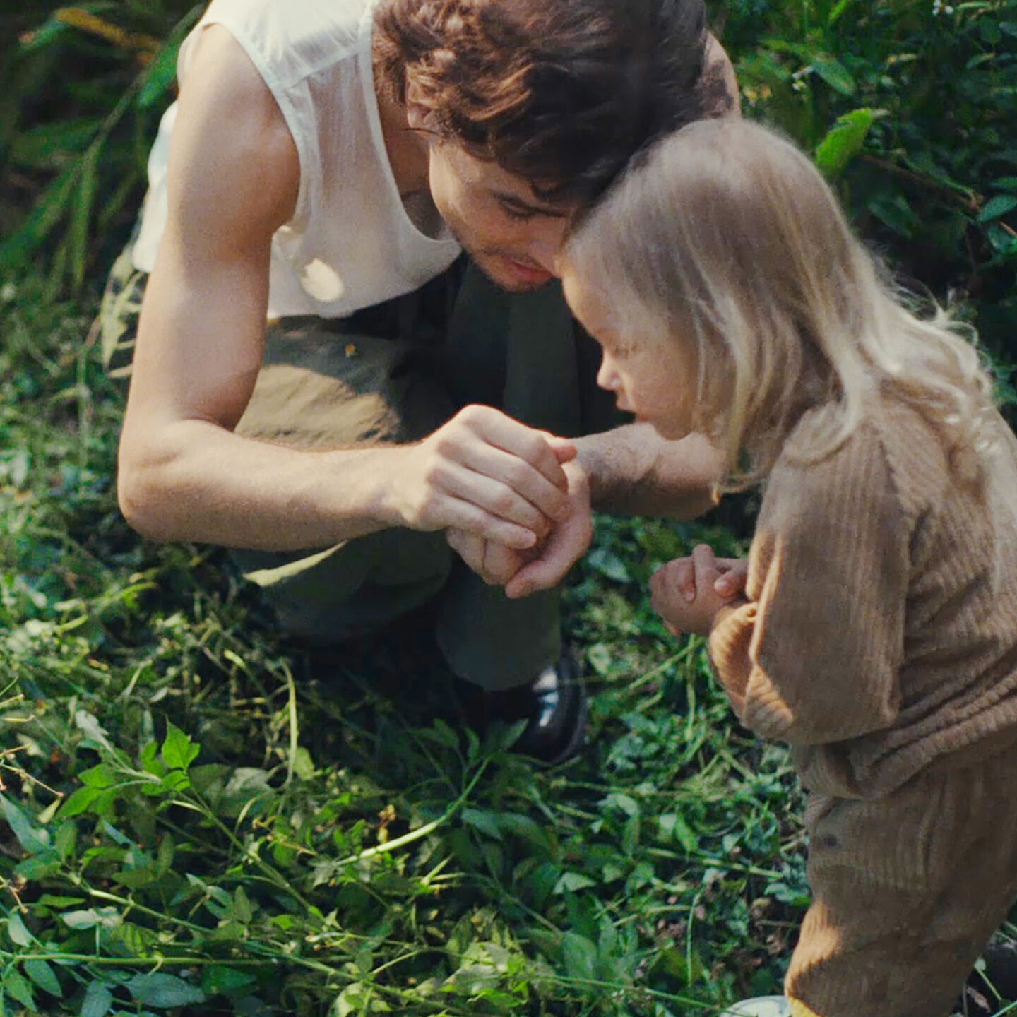生い茂った未開の庭にしゃがみこむ父親と娘。 父親は、手の中にいる捕まえた何かを娘に見せている。