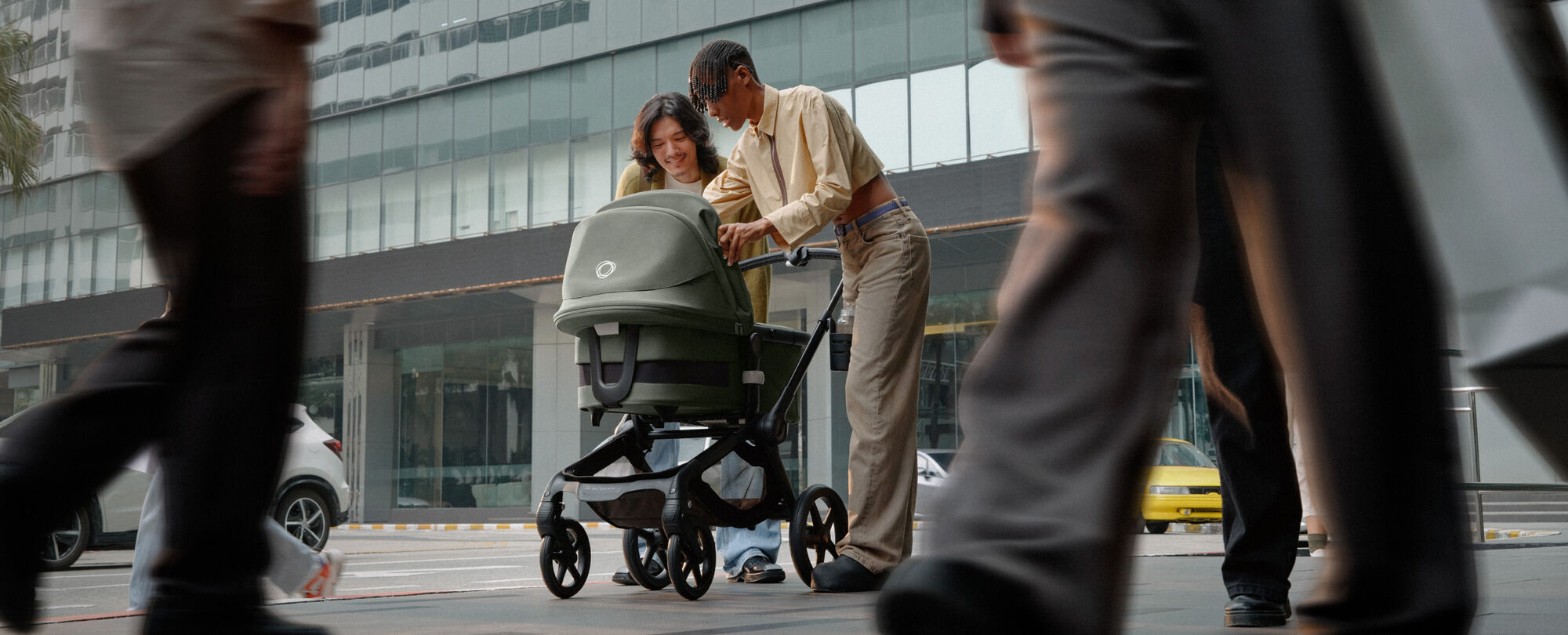 Dos padres están en medio de una calle concurrida y sonríen a su bebé dentro de un carrito Bugaboo Fox 5.