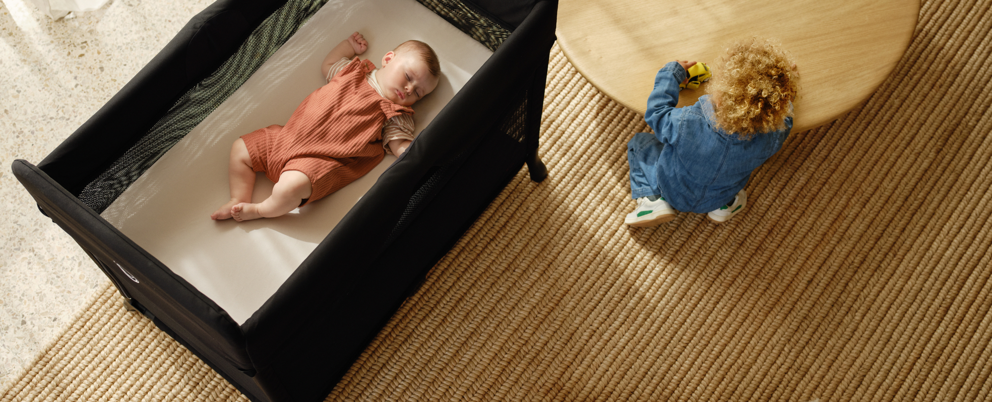 부가부 스타더스트 접이식 아기 침대 안에서 평화롭게 낮잠을 자고 있는 아이를 위에서 바라본 모습. 접이식 아기 침대 옆에는 어린 아이가 낮은 테이블 앞에서 놀고 있습니다.