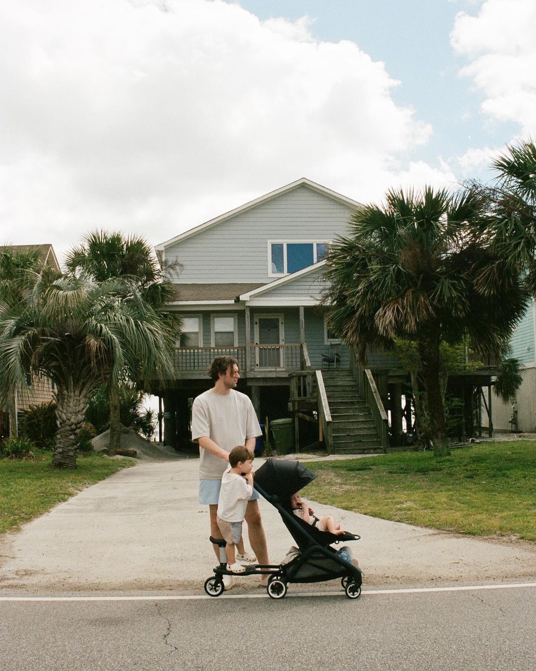 Un papá con su bebé en una Bugaboo Butterfly y su niño mayor en un patinete acoplado. Están frente a una residencia veraniega.