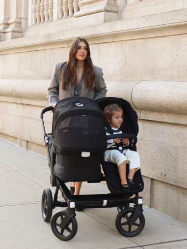 A stylish mom walks with her kids in a Bugaboo Donkey 5. Her daughter sits facing outward, while her baby in the bassinet faces her.