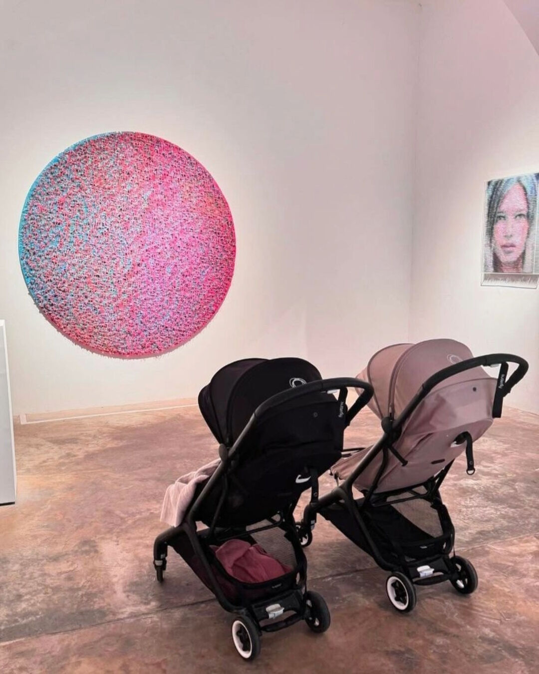 Dos sillas de paseo Bugaboo Butterfly lado a lado en una galería de arte, frente a una colorida obra artística circular y una escultura. 