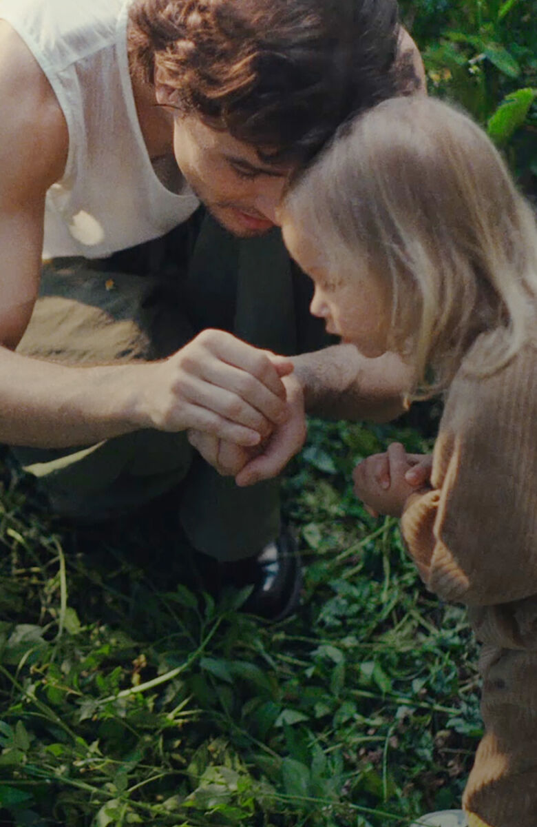 Un père et sa fille sont accroupis dans un jardin sauvage envahi de végétation. Il lui montre ce qu'il a dans ses mains.
