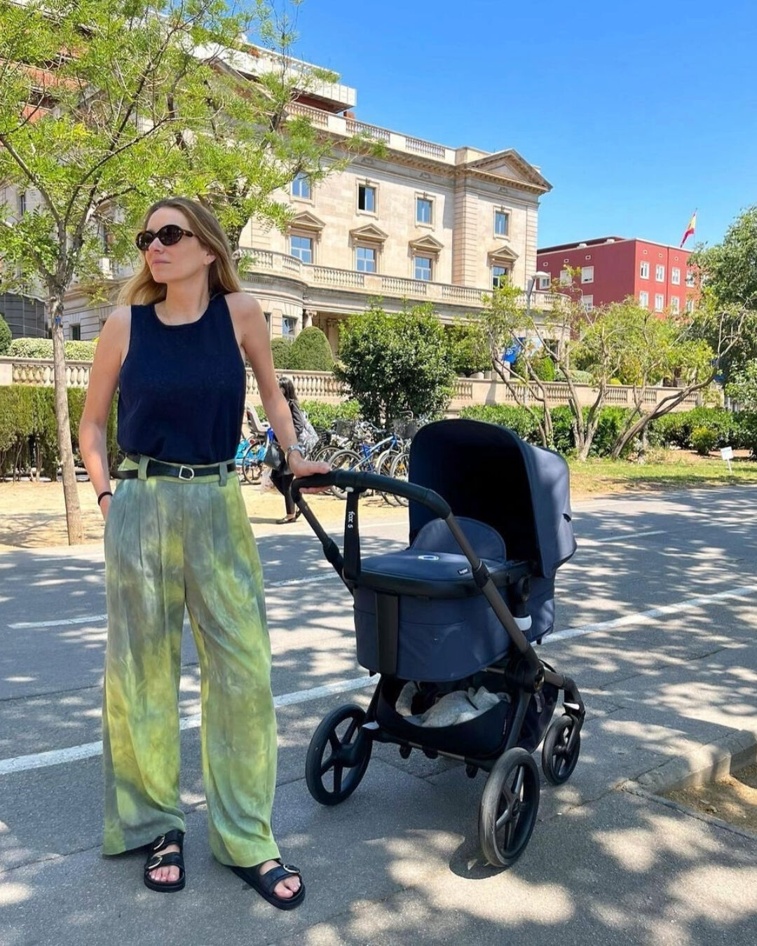 A mom poses next to her baby in a Bugaboo Fox 5. They're in an outdoor area with trees and European-style buildings in the background.