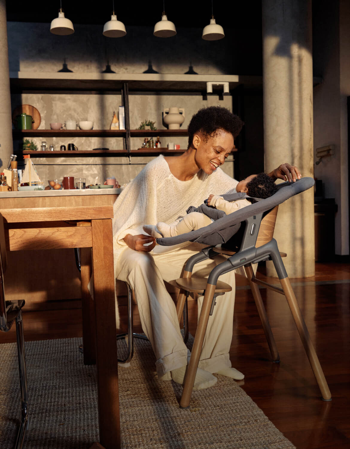 Una mamma in abiti casual gioca con il suo bambino su una sedia evolutiva Bugaboo Giraffe. Sono in una cucina moderna e illuminata da una luce calda.