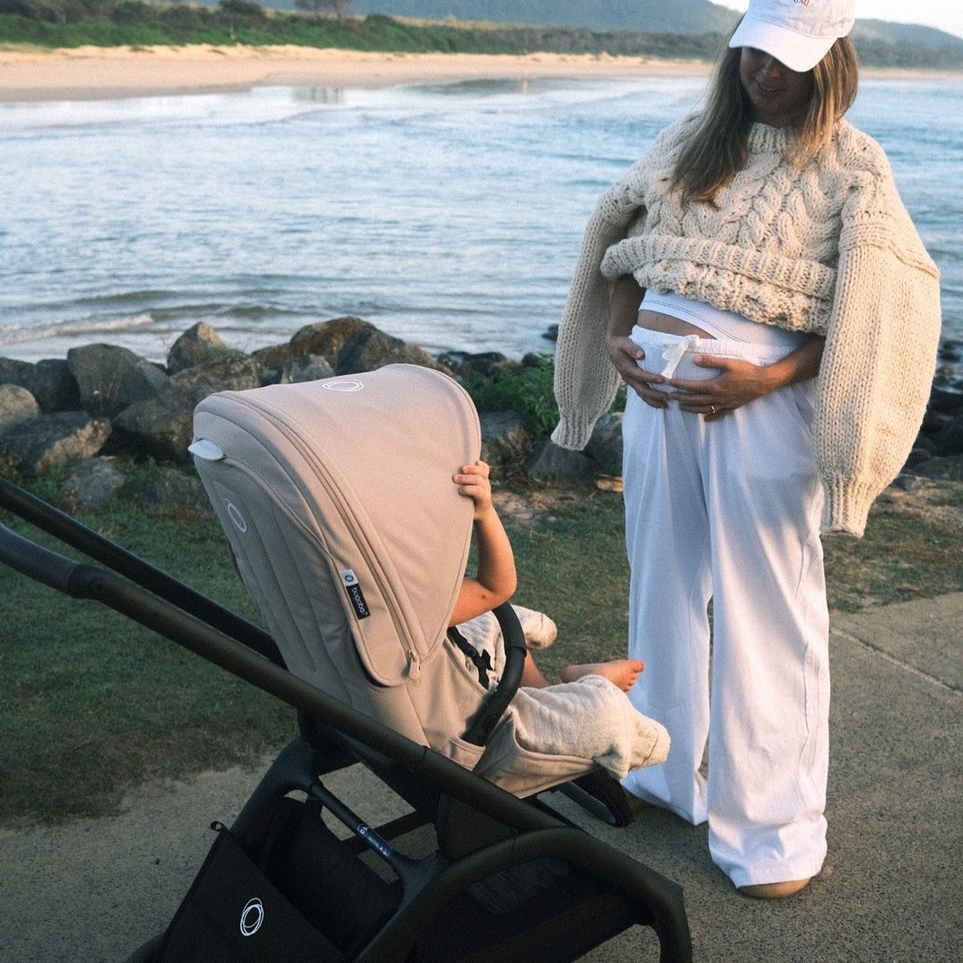 A sporty, pregnant mom smiles at her baby in a Bugaboo Dragonfly. They are on a rocky beach with the sea in the background.