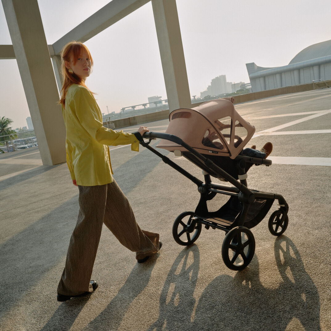 Una mamá mira a la cámara mientras pasea con su bebé en un Bugaboo Fox 5. El carrito tiene una capota ventilada en color rosa.