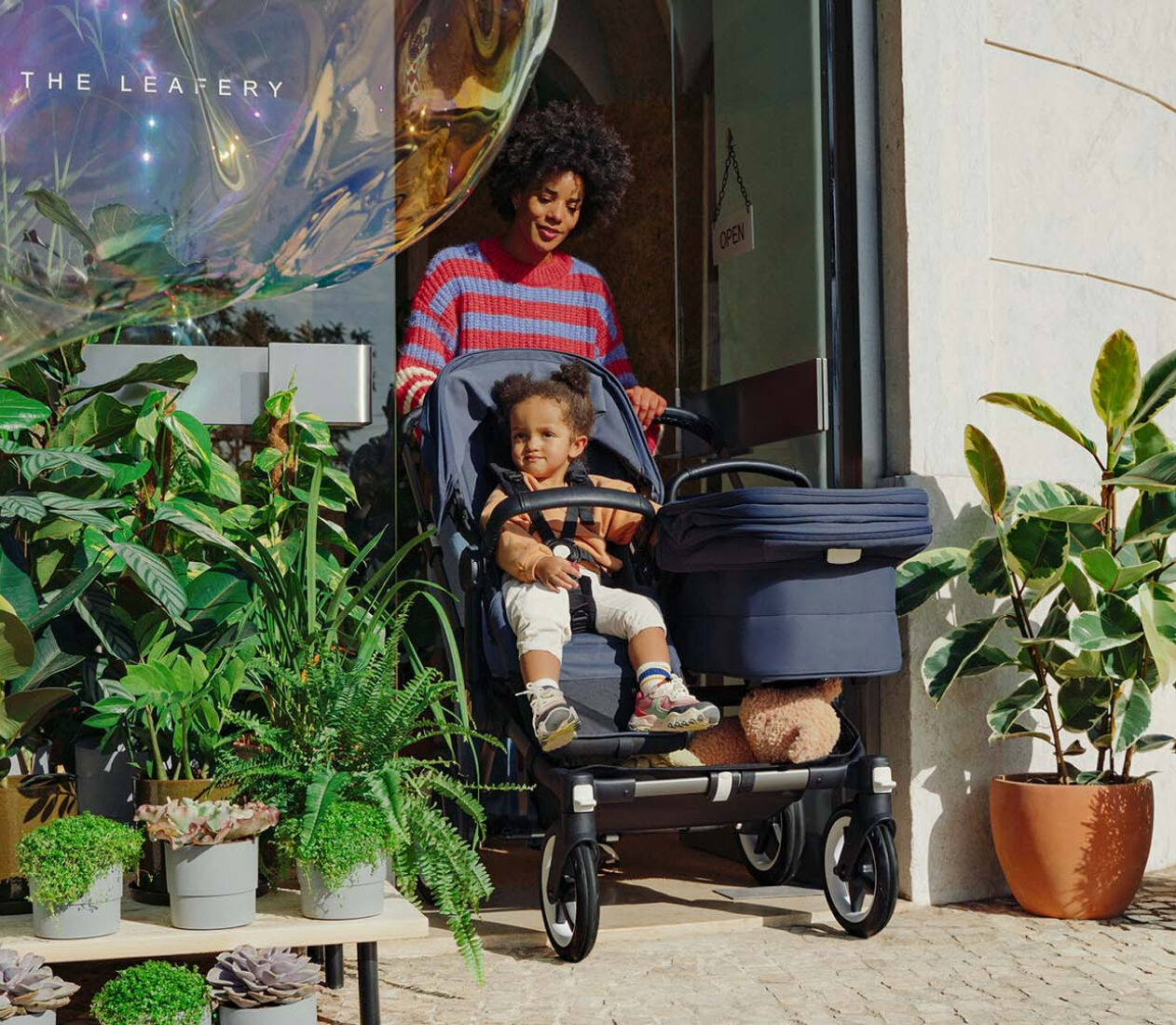 A mom exits a store, pushing a Bugaboo Donkey 5 Duo with a seat and a carrycot. A toddler sits in the seat. The store's entrance is surrounded with greenery.