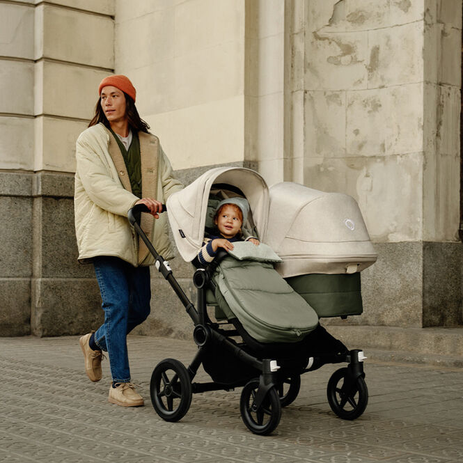 A father walks with a Bugaboo Donkey 5 Duo with a seat and a bassinet. His toddler sits on the left seat inside a footmuff.