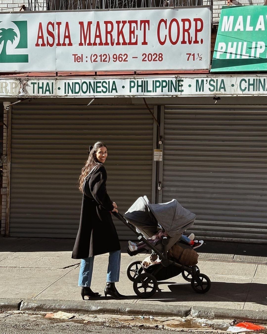 A mom walks with her two kids in a street in Southeast Asia. The kids sit in a Bugaboo Donkey 5 with the canopies extended. Behind them is a store with the sign 