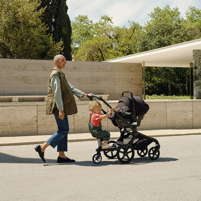 子どもたちとお散歩する母親。 彼女の赤ちゃんは、カーシートを装着したバガブー フォックス 5に乗っている。 年上の子どもはホイールボードに乗っている。