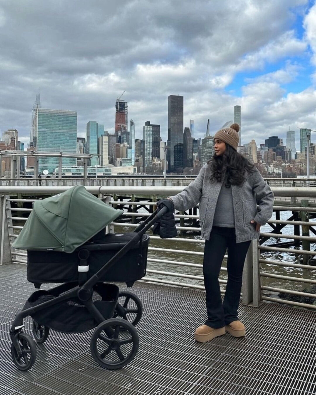 A warmly-dressed mom and her baby in a Bugaboo Fox 5. They stand on a promenade, looking out to a city skyline.