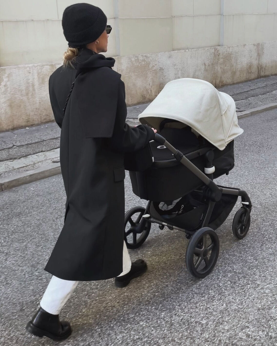 Eine stylische Mutter geht mit ihrem Baby spazieren, das in der Liegewanne eines Bugaboo Fox 5 mit ausgefahrenem Sonnendach schläft.