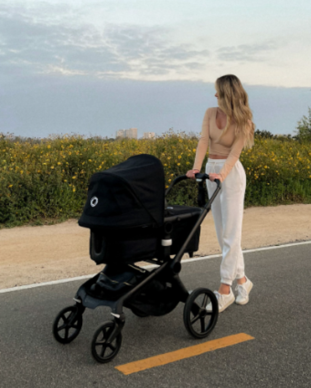 A mom walks her baby in a Bugaboo Fox 5 on a paved road. In the background are a flower field and a partly cloudy sky.